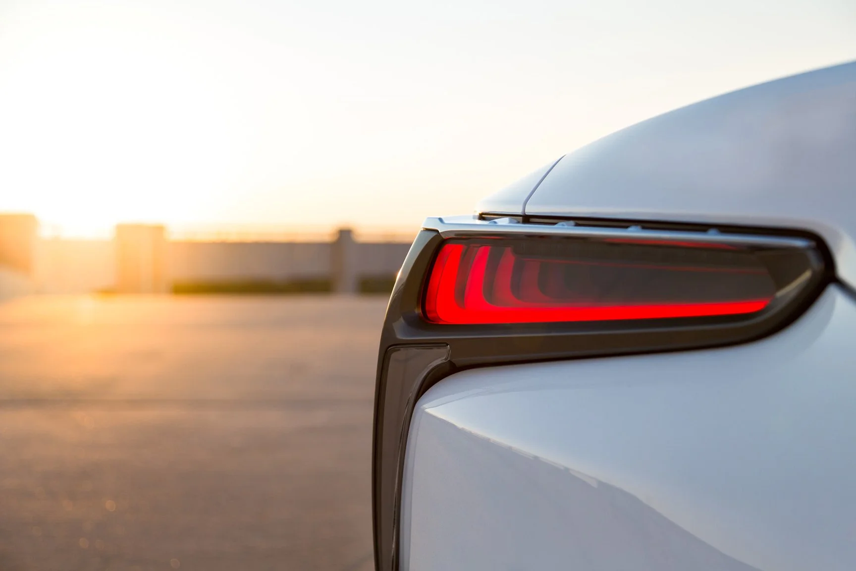 Close-up of the rear right corner of a white car, showing a modern red tail light, with a blurred sunset background.