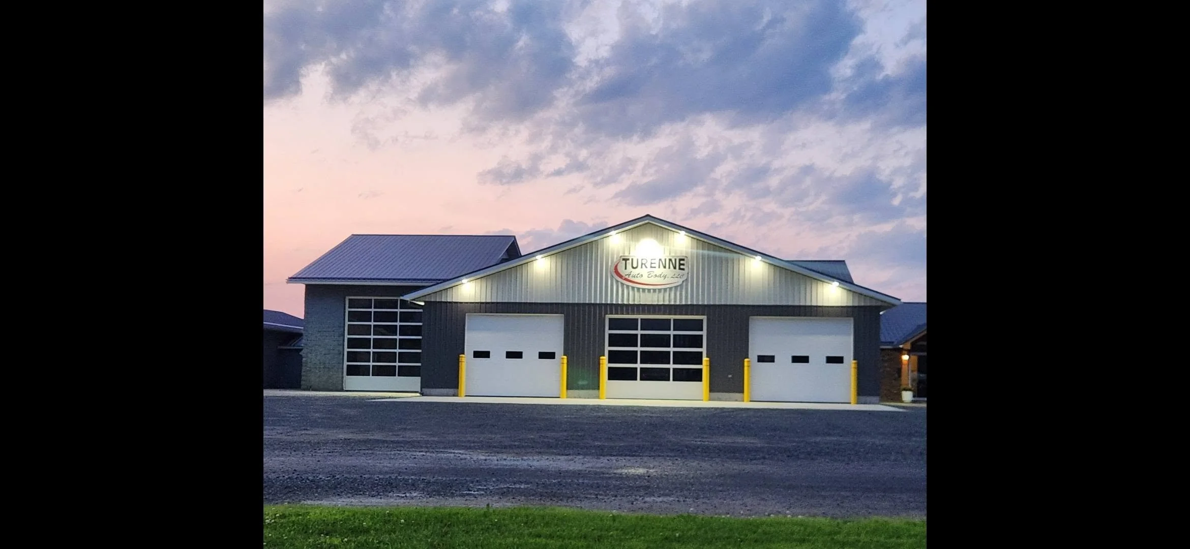 A commercial garage building with four large garage doors, three yellow parking barriers, and a sign that reads "Turenne Auto Body LLC" on the front. The building is illuminated by exterior lights against a sunset sky with scattered clouds.