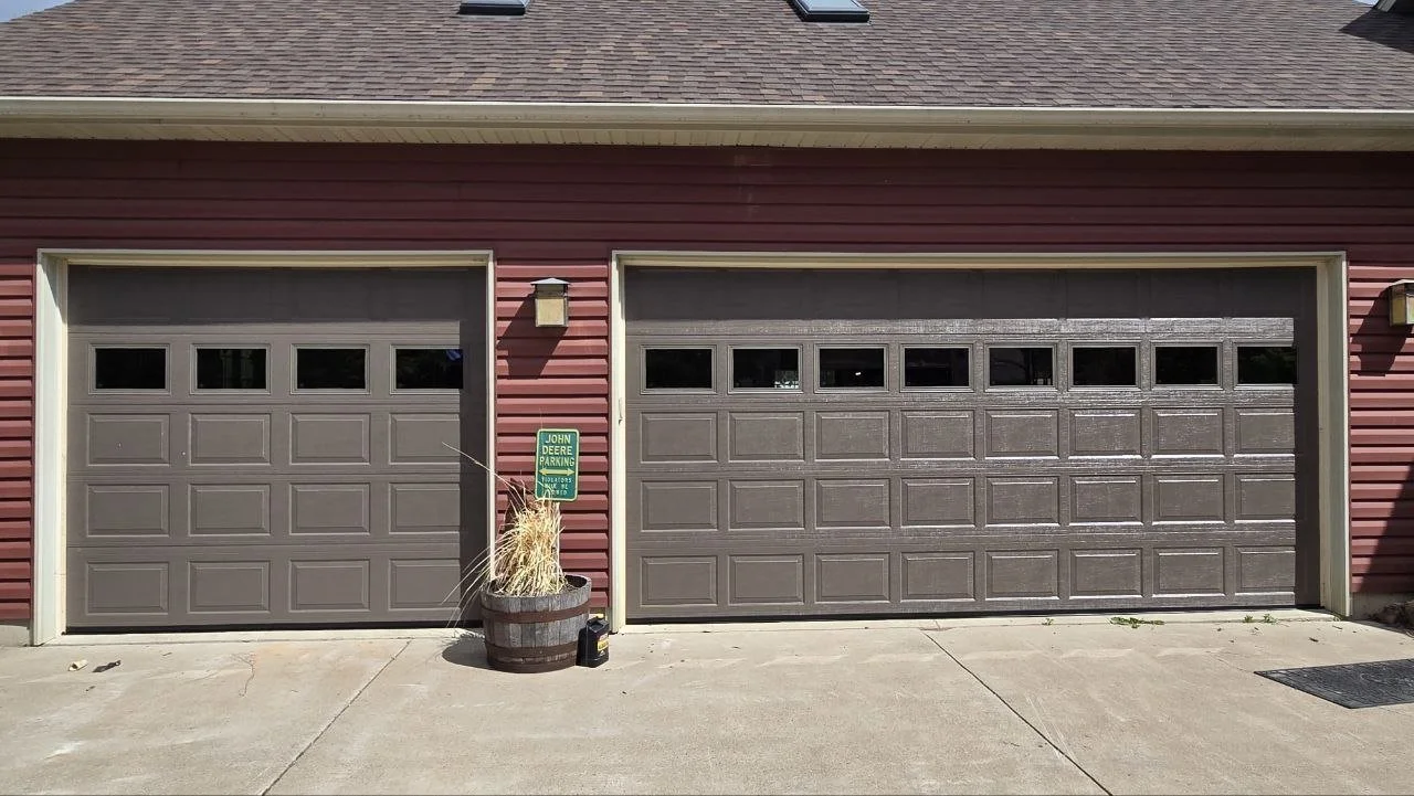 Two sectional garage doors on a red house, with a green 'John Deere Parking' sign, a potted plant, and a small black container in front.