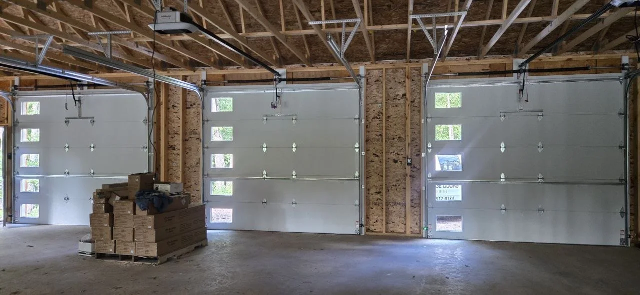 Three white garage doors in an unfinished garage with exposed wood framing, some construction materials on a pallet in the center, and natural light coming through small windows in the doors.