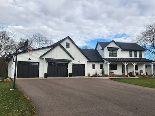A large white house with black roofing and garage doors, a front porch with seating, and a spacious driveway, set in a suburban neighborhood with trees and cloudy sky