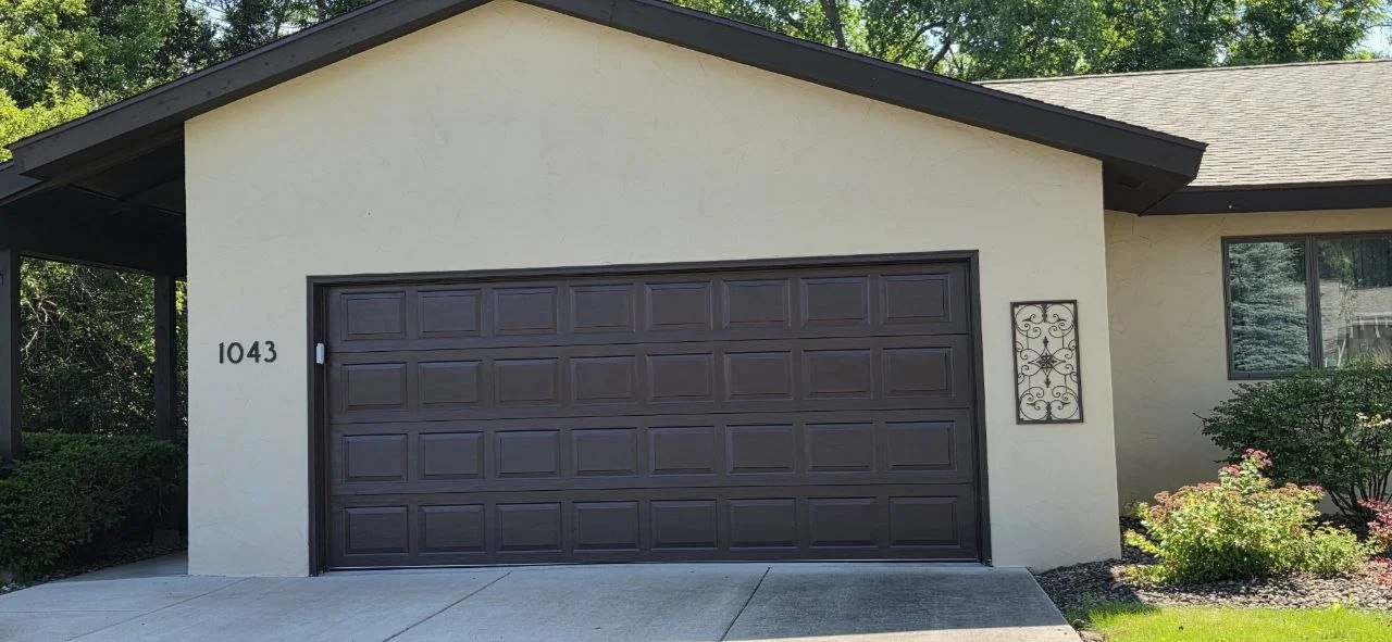 Front view of a house showing a closed dark brown garage door, house number 1043 on a beige wall, a window, and a flower garden with bushes and flowering plants.