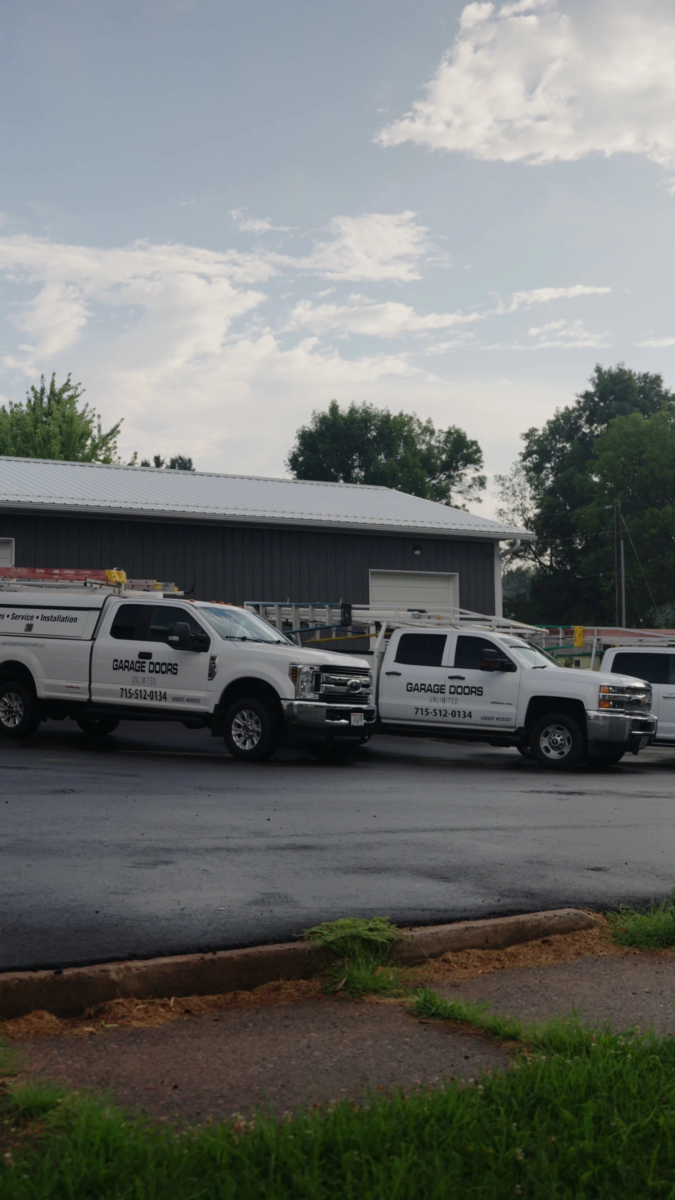 Two white service trucks parked in front of a dark gray building with a metal roof, with a partly cloudy sky and trees in the background.