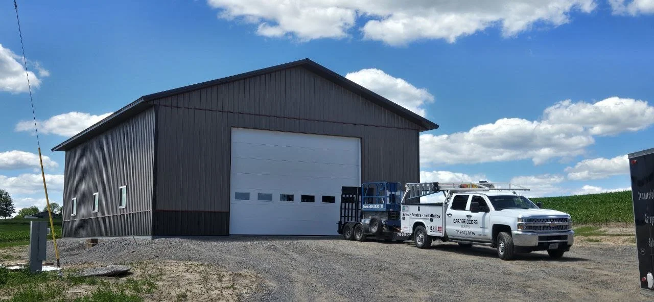 Large metal barn with a white garage door, on a gravel lot with two service trucks parked in front, under a partly cloudy sky.