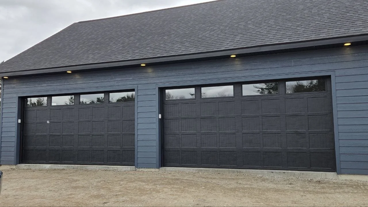 A blue garage with two black garage doors and small rectangular windows at the top, under a sloped roof with small exterior lights underneath.