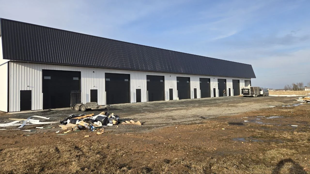 Long industrial building with black doors and a metal roof, construction debris and trucks outside, under a partly cloudy sky.
