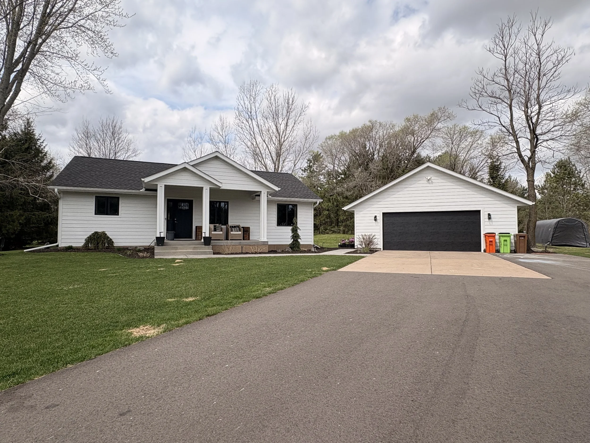 White house and garage with dark roof, surrounded by trees, with a driveway and recycling bins in front.