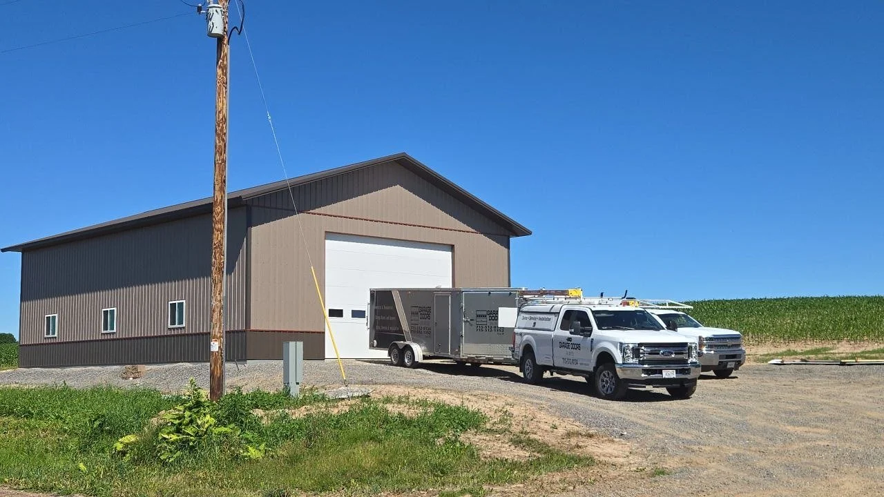A rural scene with a large metal building, a utility pole, and two service trucks parked in front on a gravel driveway, with green fields and a clear blue sky in the background.
