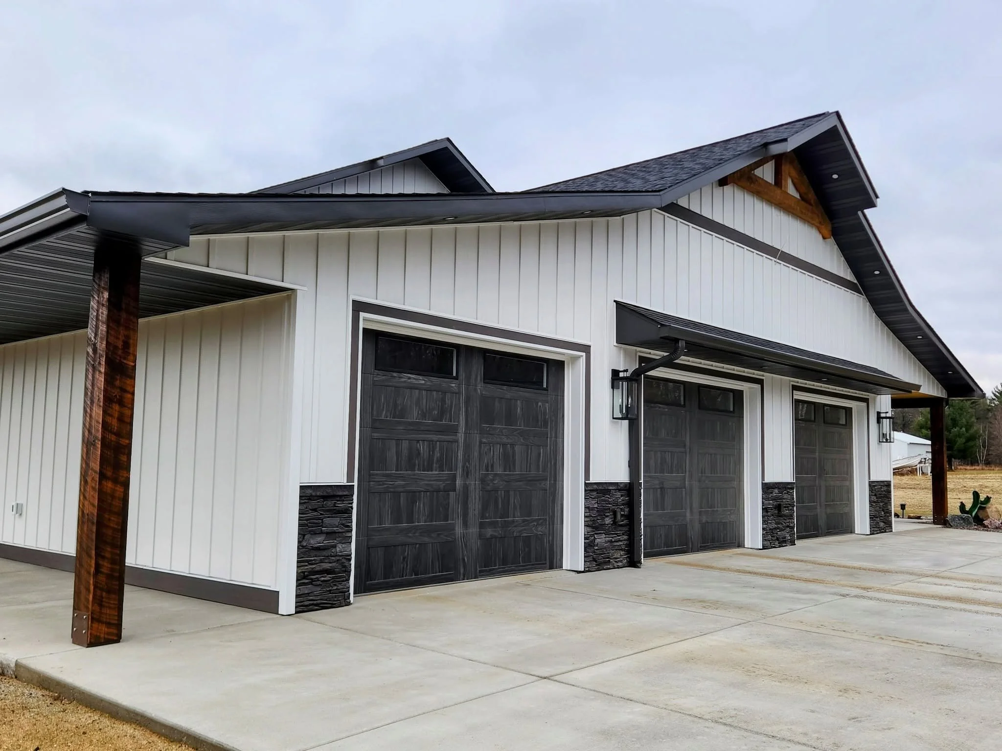 A modern residential house with a dark gray garage door, white siding, and stone accents. The house features a sloped roof with exposed wooden beams and a concrete driveway. There is a lamppost and a wooden post near the driveway, with an overcast sk