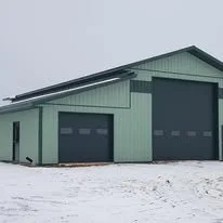 A green metal building with two large garage doors and a smaller door, set in a snowy landscape under a gray sky.