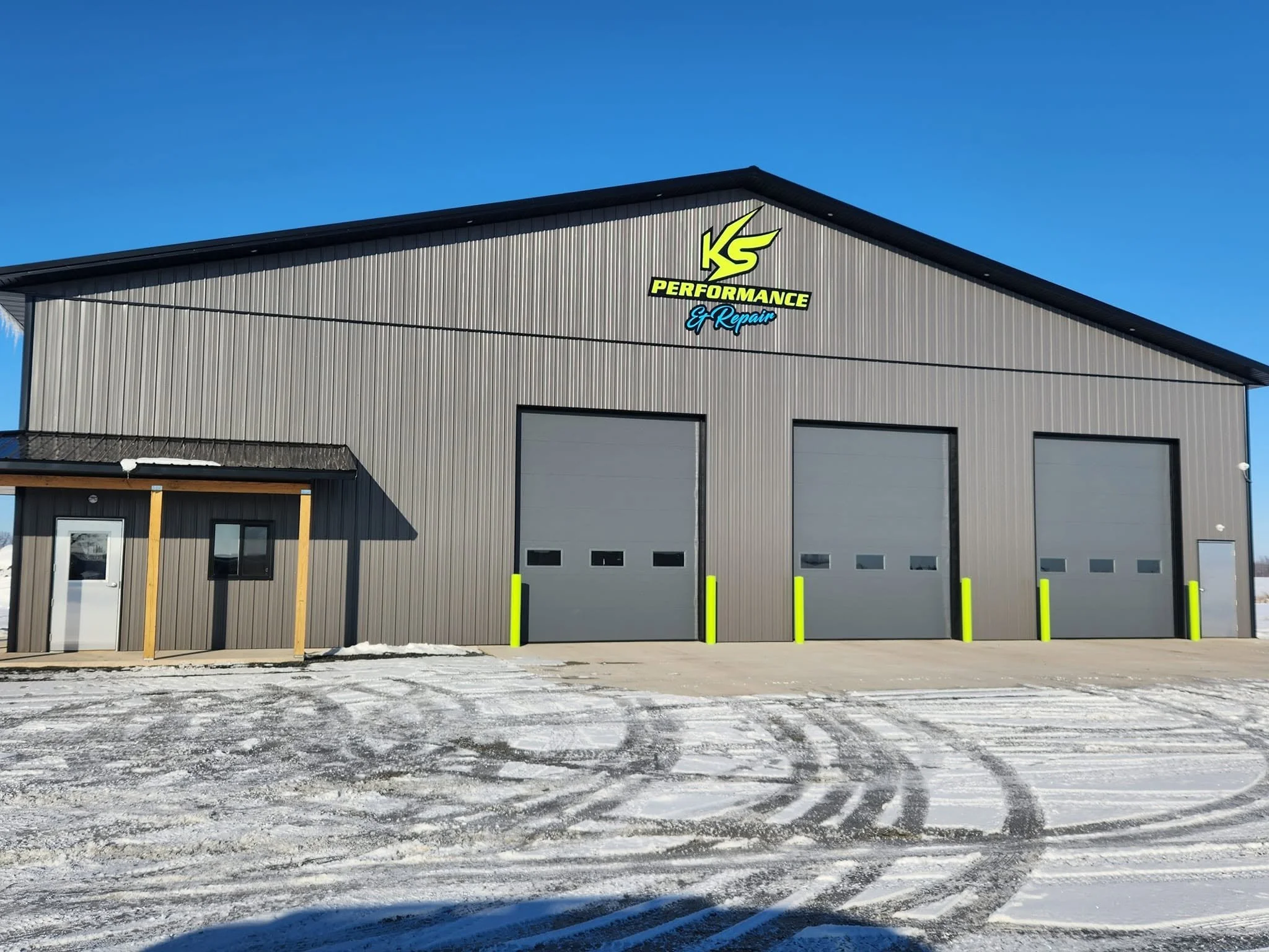 Front view of a large industrial building with gray metal siding and three garage doors, with a parking lot in front that has patches of snow and ice, under a clear blue sky. The building features signage for KS Performance & Repair.