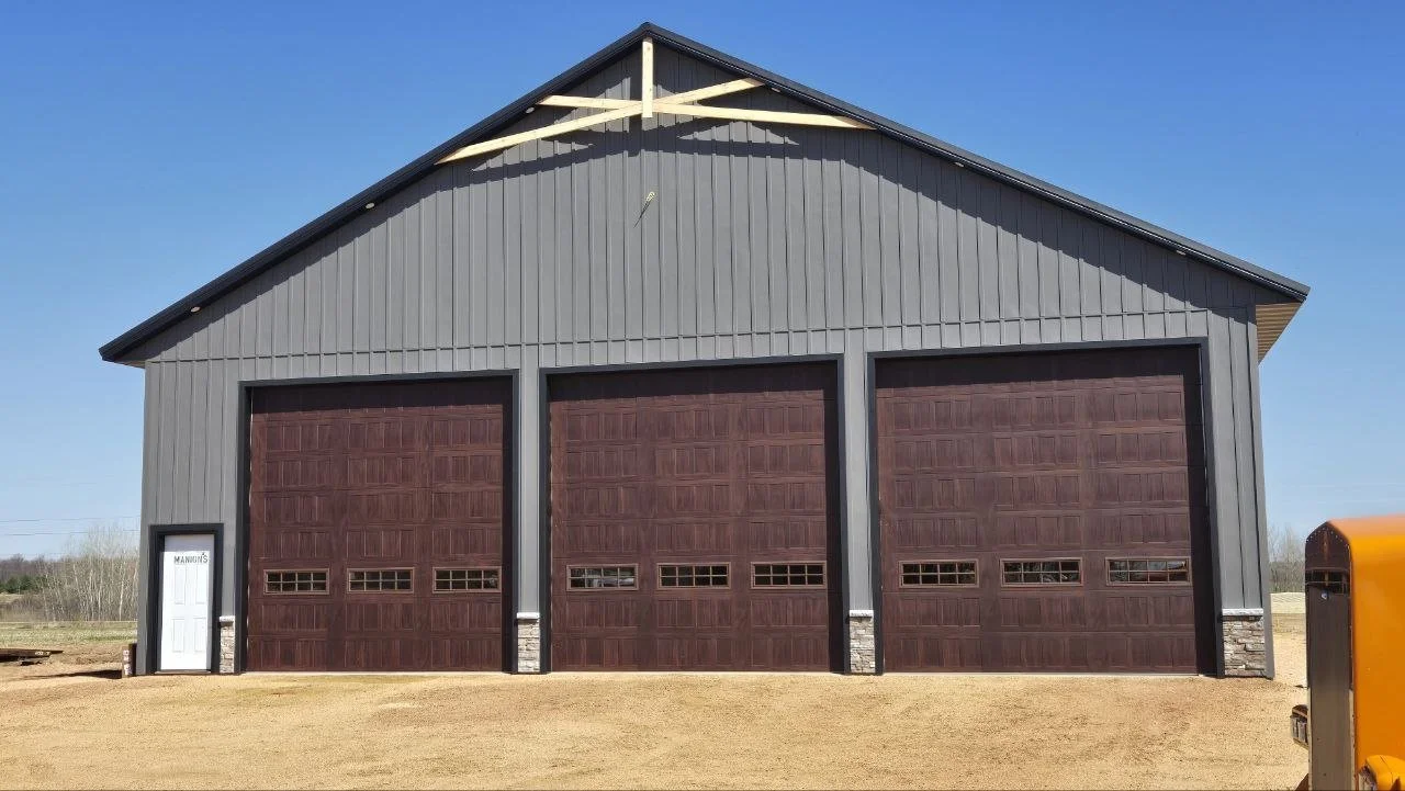 Large gray metal building with three brown garage doors and a small white door labeled "MANU'S" on the left, set against a clear blue sky and dirt ground.