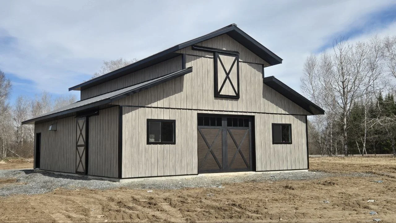 A large barn with gray wooden siding, black trim, and a metal roof, situated on a partly cleared piece of land with trees in the background.