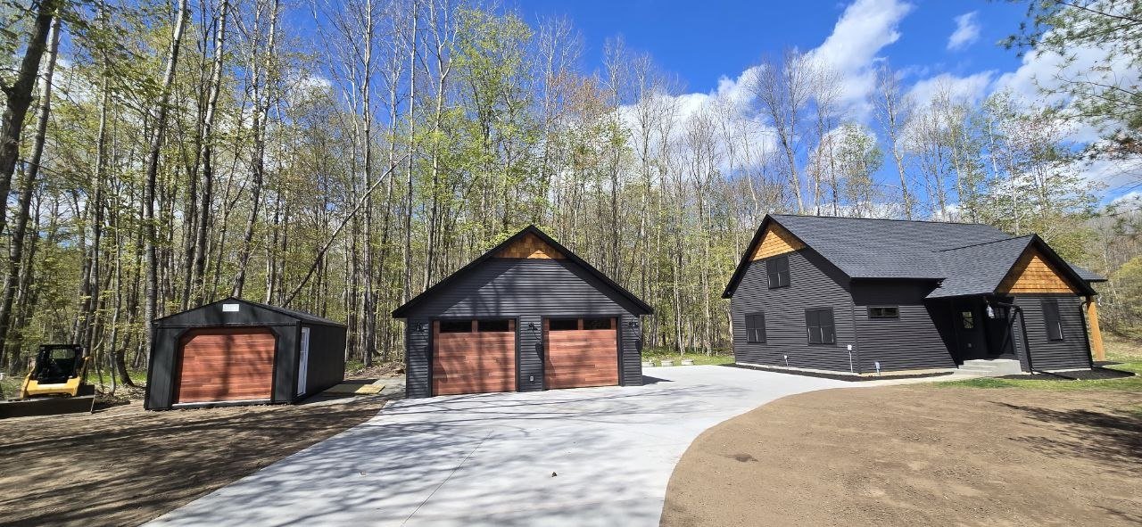Modern black house with brown garage doors, surrounded by trees, with a concrete driveway and a separate black garage with a brown door on the left.