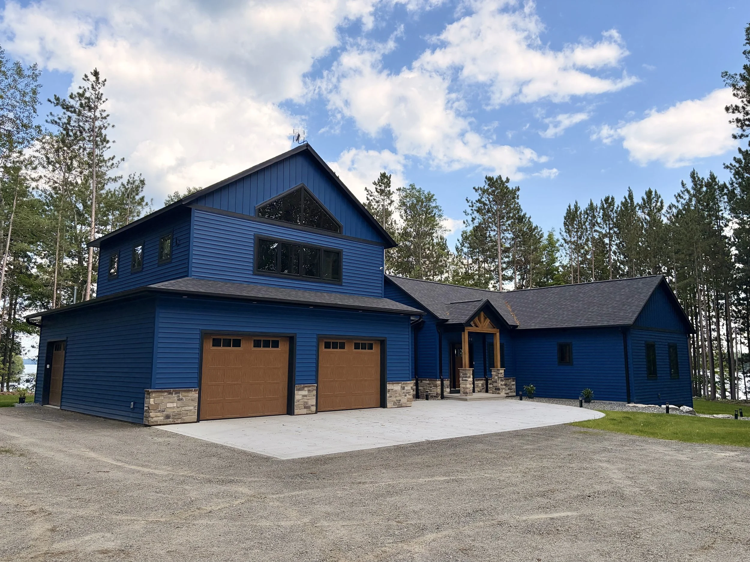 A modern blue house with a stone accent on the base, large garage doors, and a surrounding gravel driveway, set against a wooded background with tall trees and a partly cloudy sky.