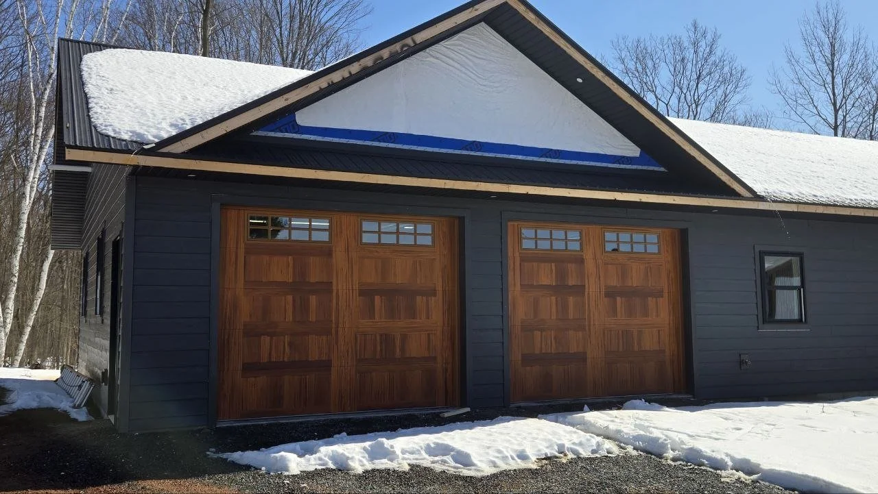 A house with two wooden garage doors, dark blue siding, and a snow-covered roof during winter. Some snow is seen on the ground in front of the garage.