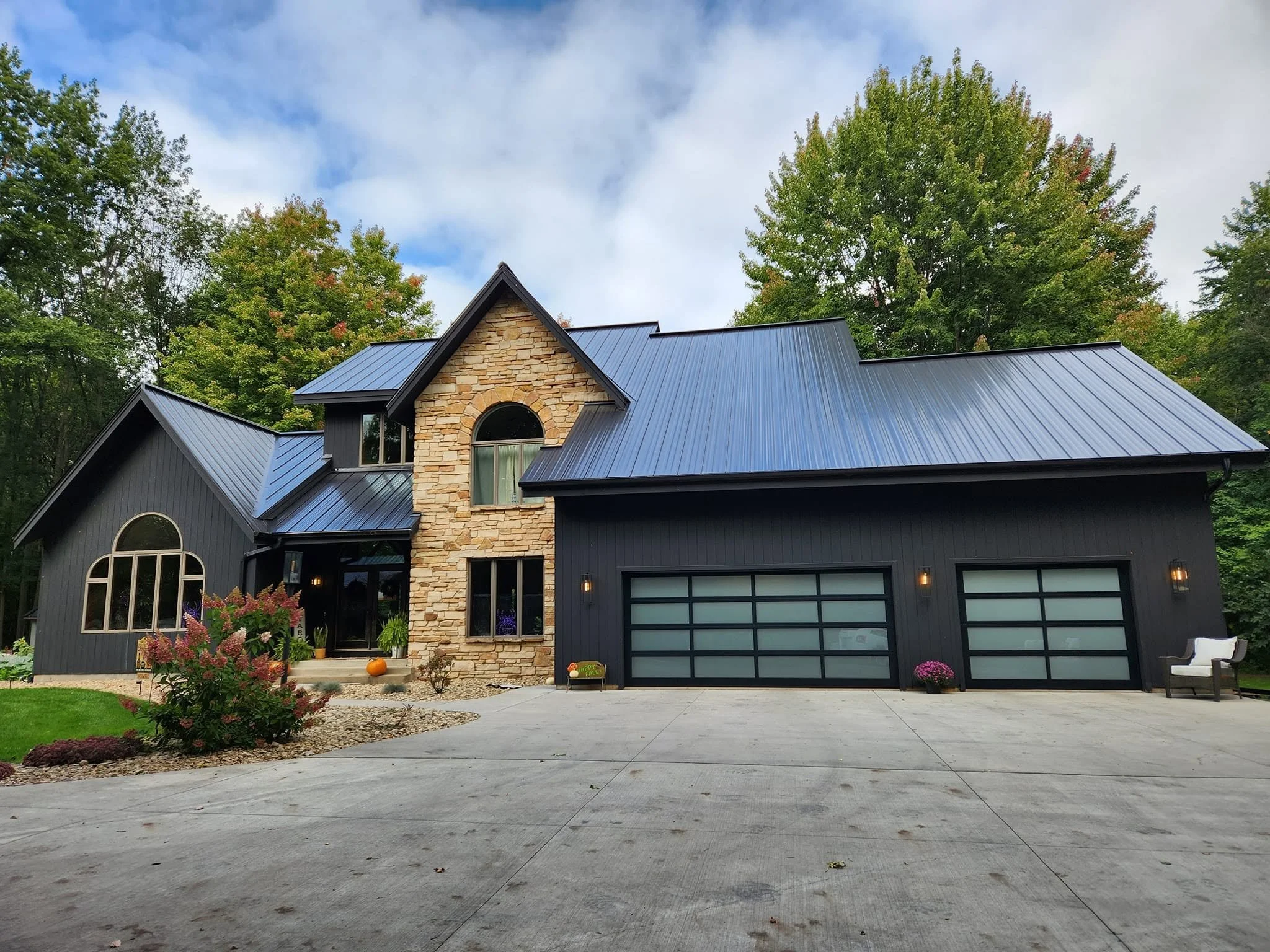 A modern house with dark gray siding and a stone front, surrounded by green trees under a partly cloudy sky, with a concrete driveway and front yard decorated with plants and pumpkins.
