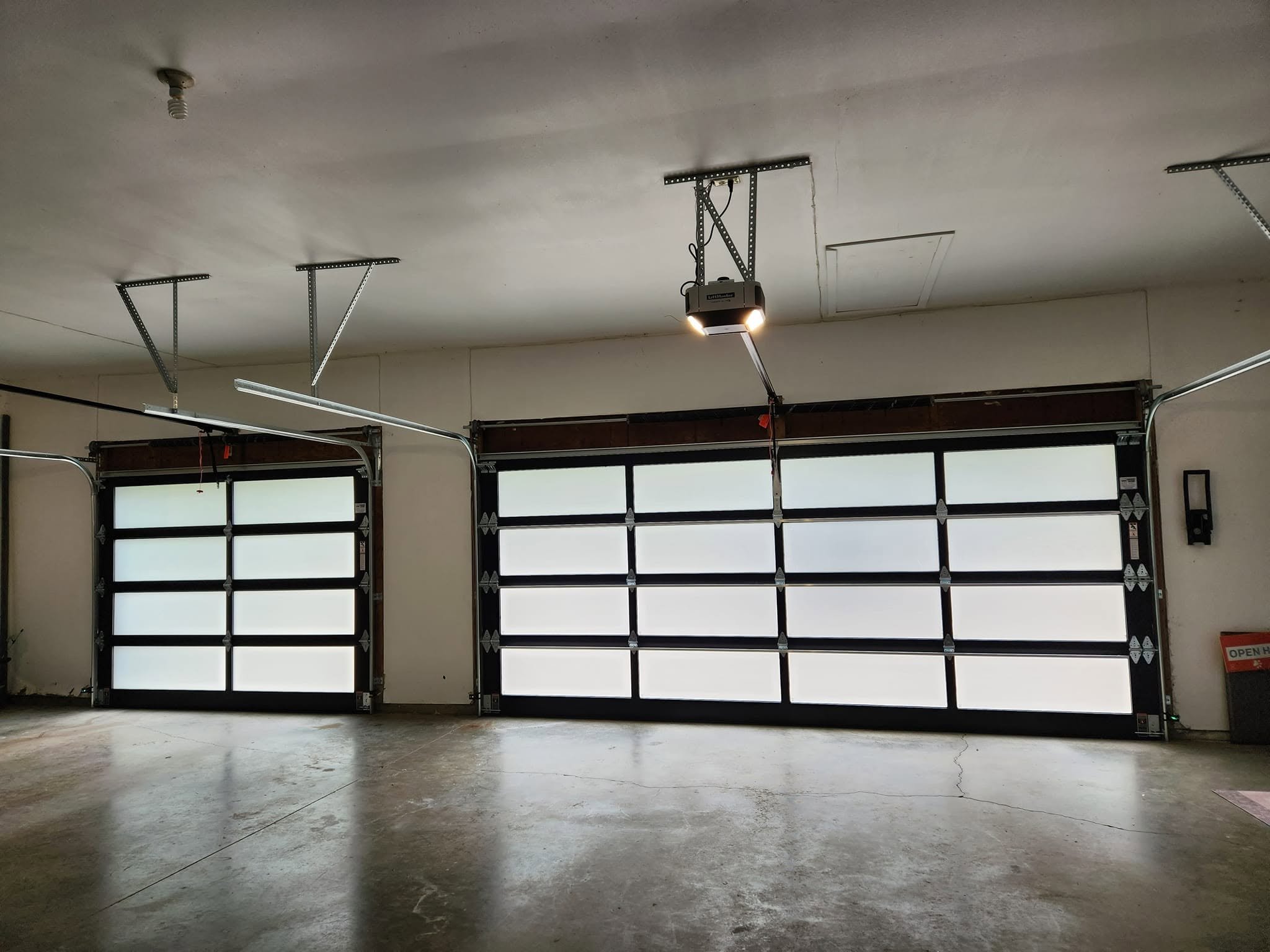 Garage with four white garage doors with small windows at the top, a garage door opener on the ceiling, and concrete floor with some cracks.