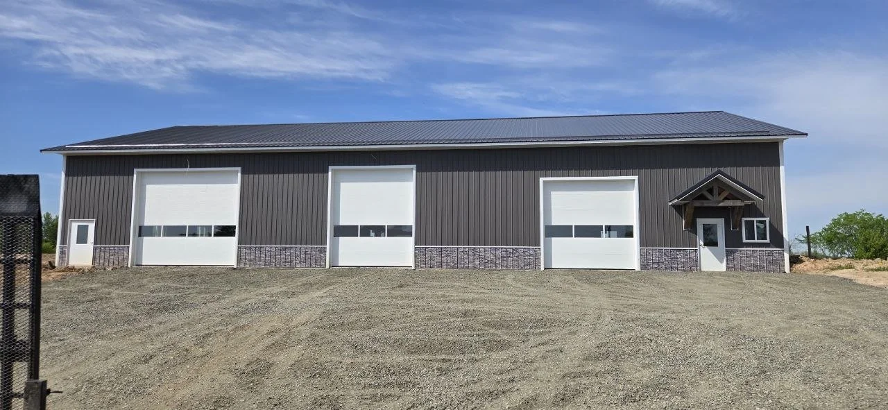 Large gray metal building with three garage doors, a small pedestrian door with a porch, and a dirt foreground.
