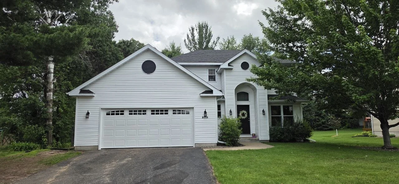 White two-story house with an attached garage and a black driveway, surrounded by green trees and grass, with cloudy skies overhead.