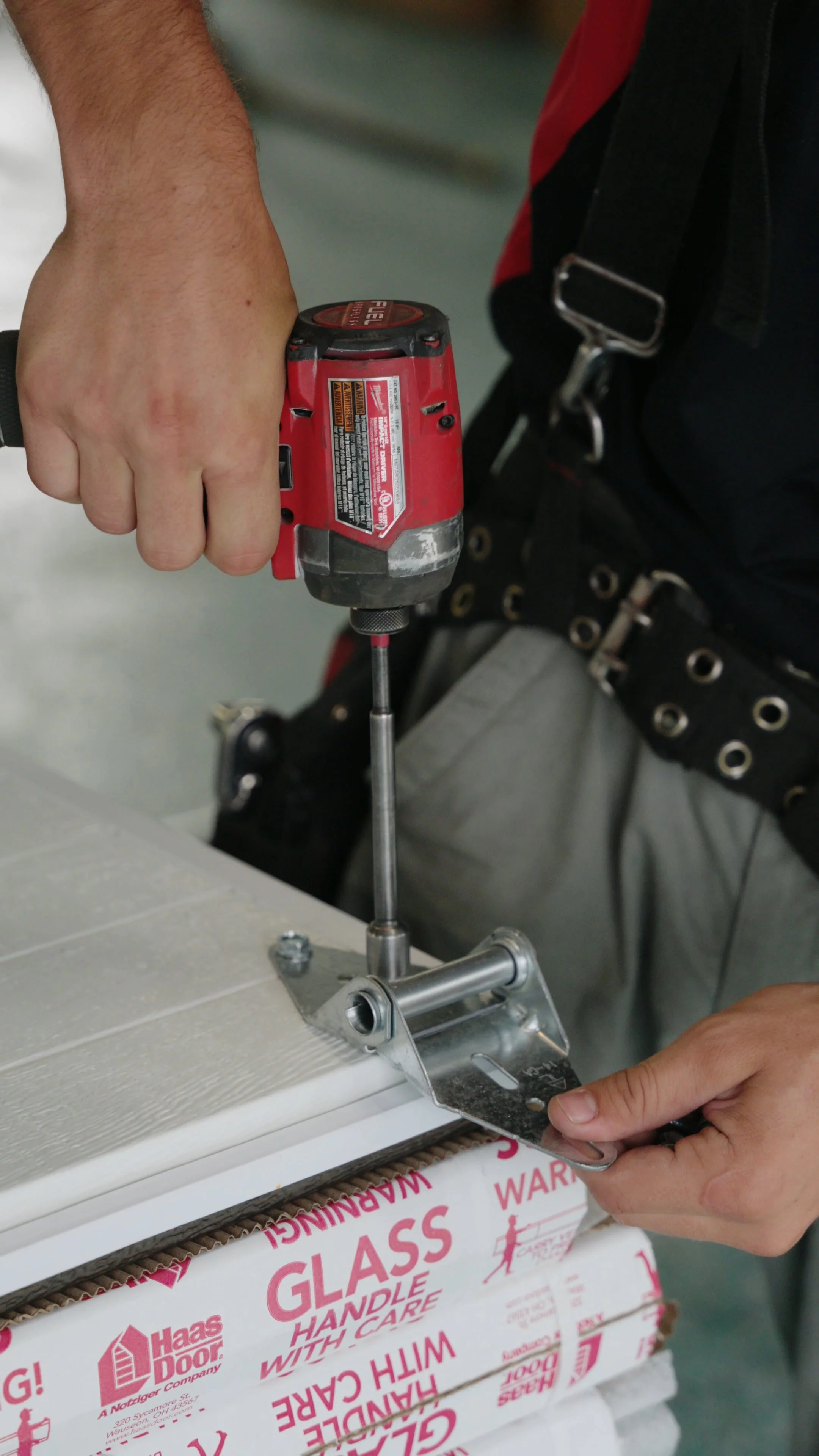A person operating a cordless power drill to fasten a metal bracket to a stack of boxes with glass handling labels, while standing in a workspace.