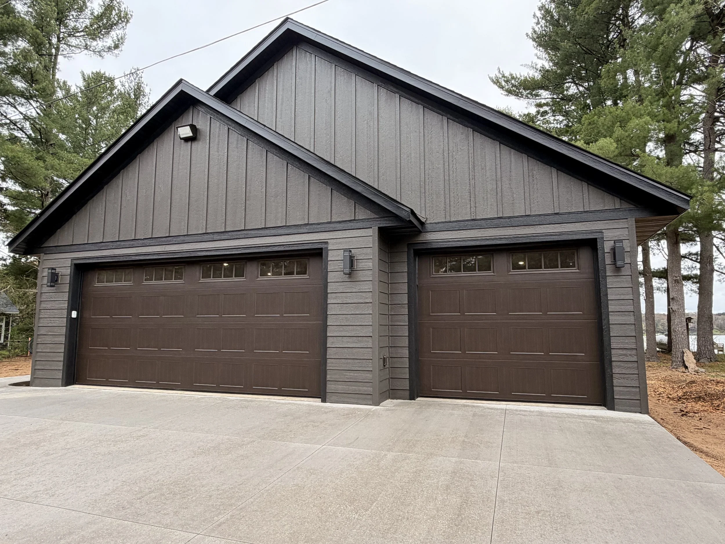 Exterior of a modern house with a dark garage door, grey wooden siding, and triangular rooflines, surrounded by trees on a cloudy day.
