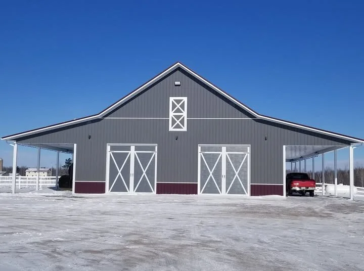 A large gray barn with white trim and two sets of white double doors, situated on snow-covered ground under a clear blue sky. A red pickup truck is parked on the right side of the barn.