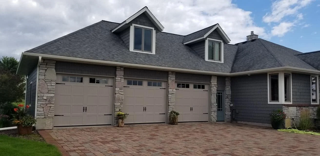 front view of a large house with three attached garage doors, gray siding, stone accents, a pitched roof with dormer windows, and a brick driveway.