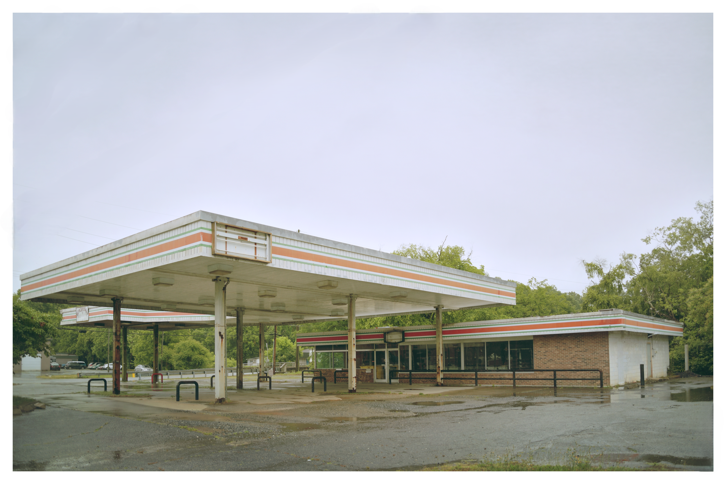 An abandoned gas station with an empty canopy, rusted support poles, and a flooded parking lot surrounded by trees and other buildings.