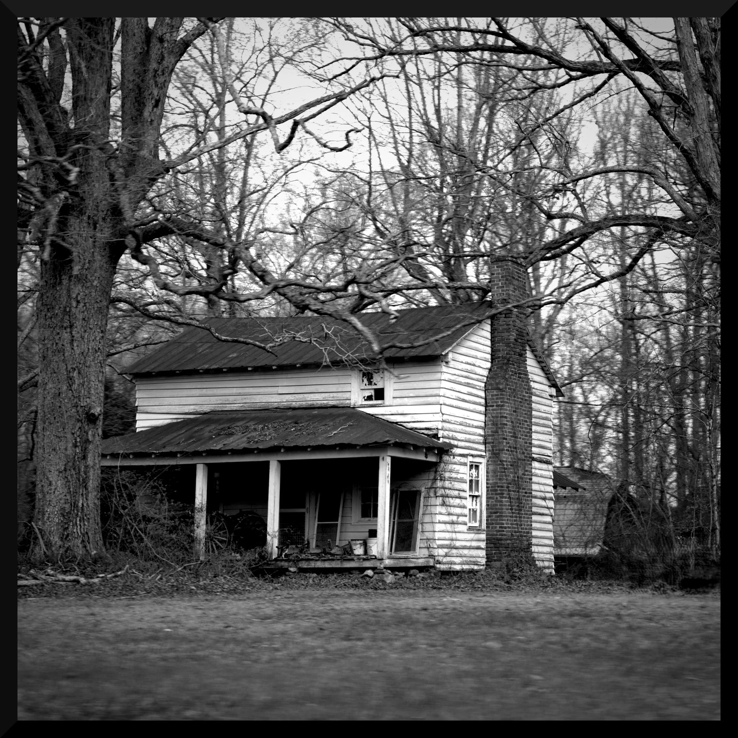 Old, abandoned house with peeling paint, broken windows, and a chimney, surrounded by leafless trees, in black and white.
