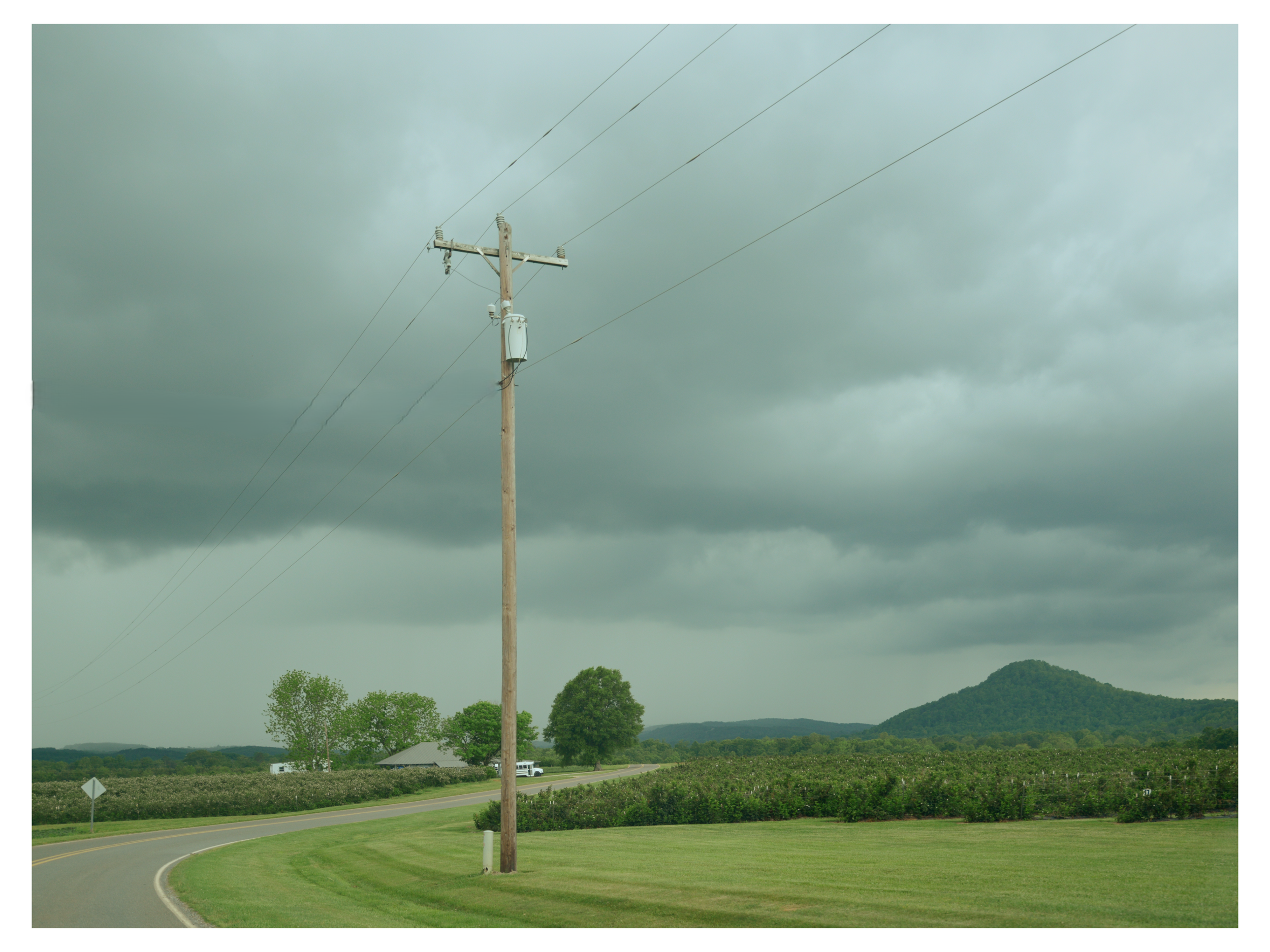 A utility pole with power lines against a cloudy sky, with green trees and rolling hills in the background.