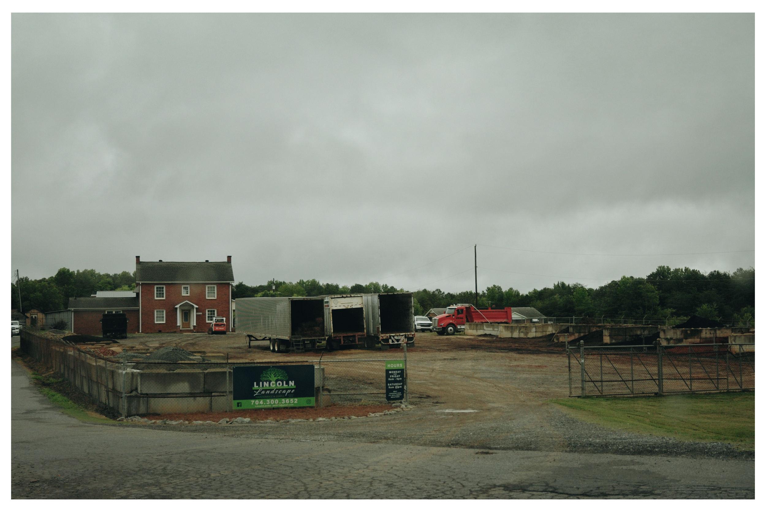 A construction site with a red house and several trucks, including an open box truck, on a cloudy day. There is a sign for Lincoln Landscape near the entrance.