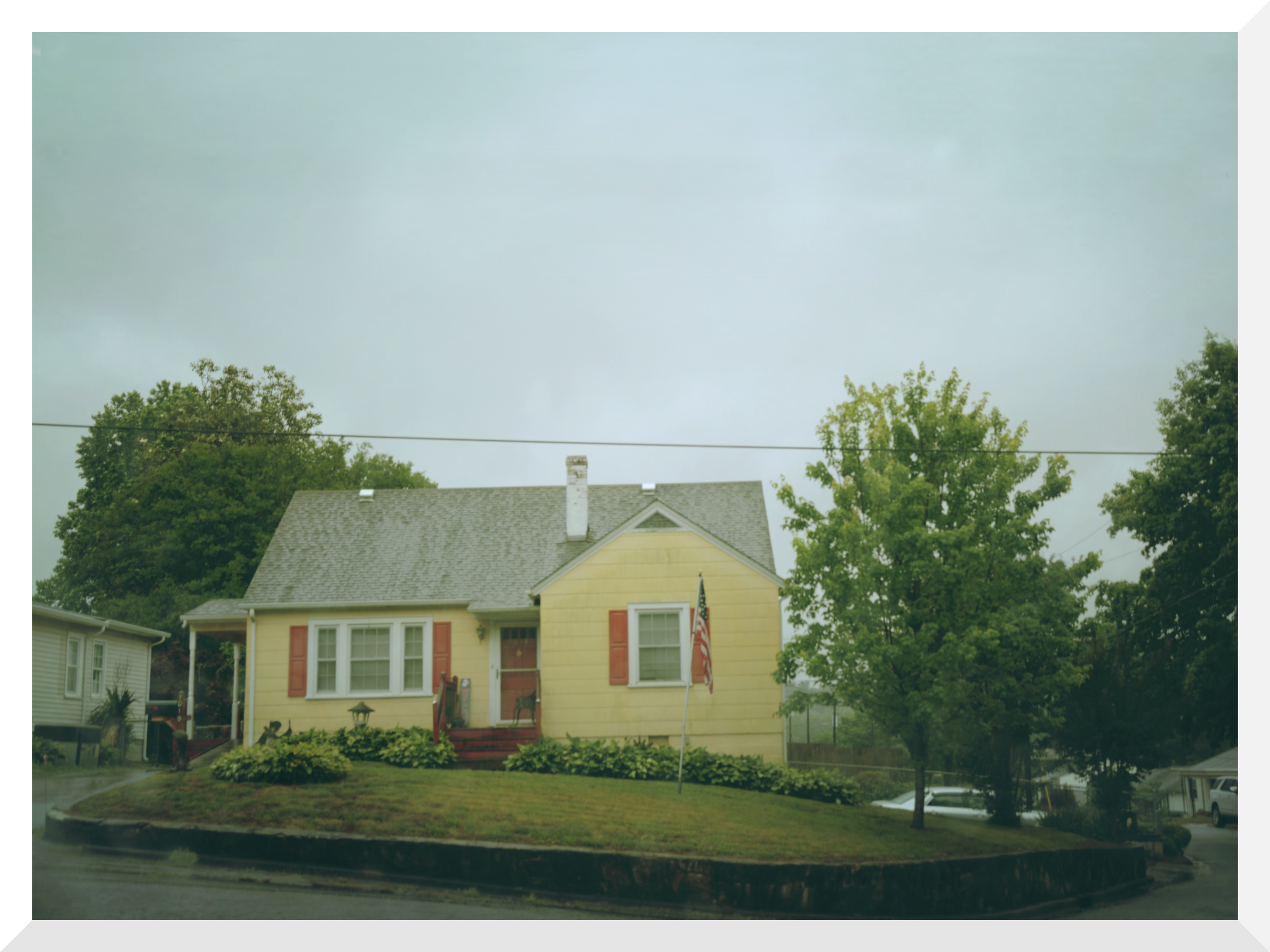 A yellow house with red shutters and white trim, featuring a small front porch with a red railing, surrounded by trees and a well-maintained lawn, with a cloudy sky overhead.
