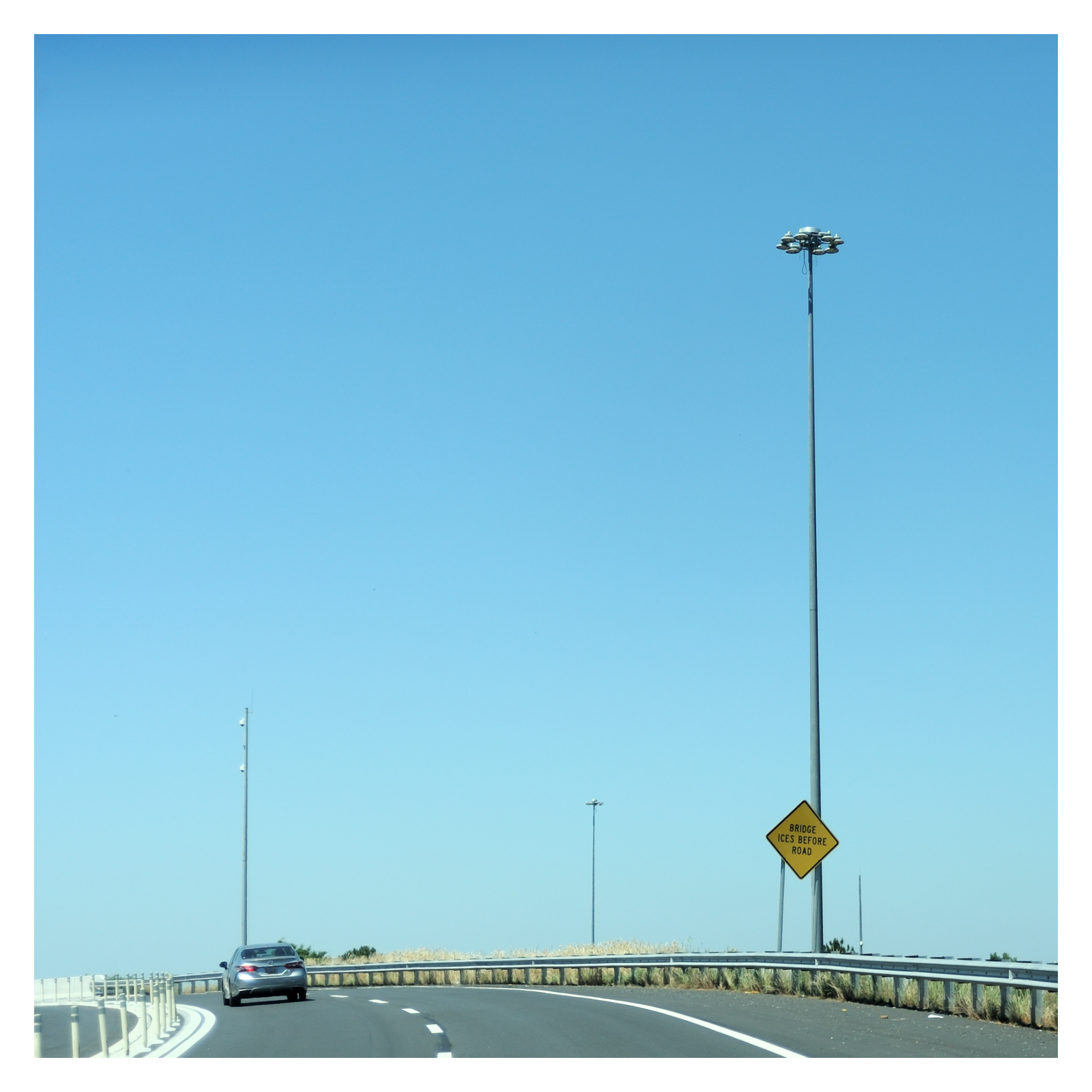 A highway scene with a single car on a curved road, multiple tall light poles with security cameras, and a yellow traffic sign that reads 'BRIDGE ICE BEFORE ROAD' under a clear blue sky.