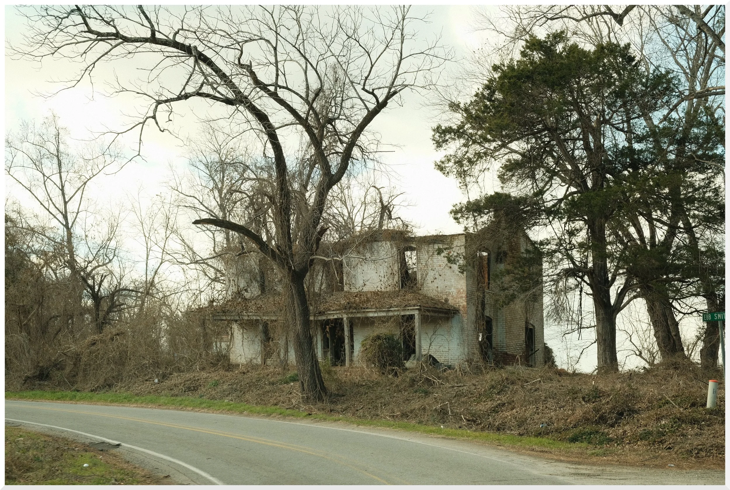 Old, abandoned house surrounded by leafless trees, located near a road, with a street sign reading 'Ebb Smith' in the lower right corner.