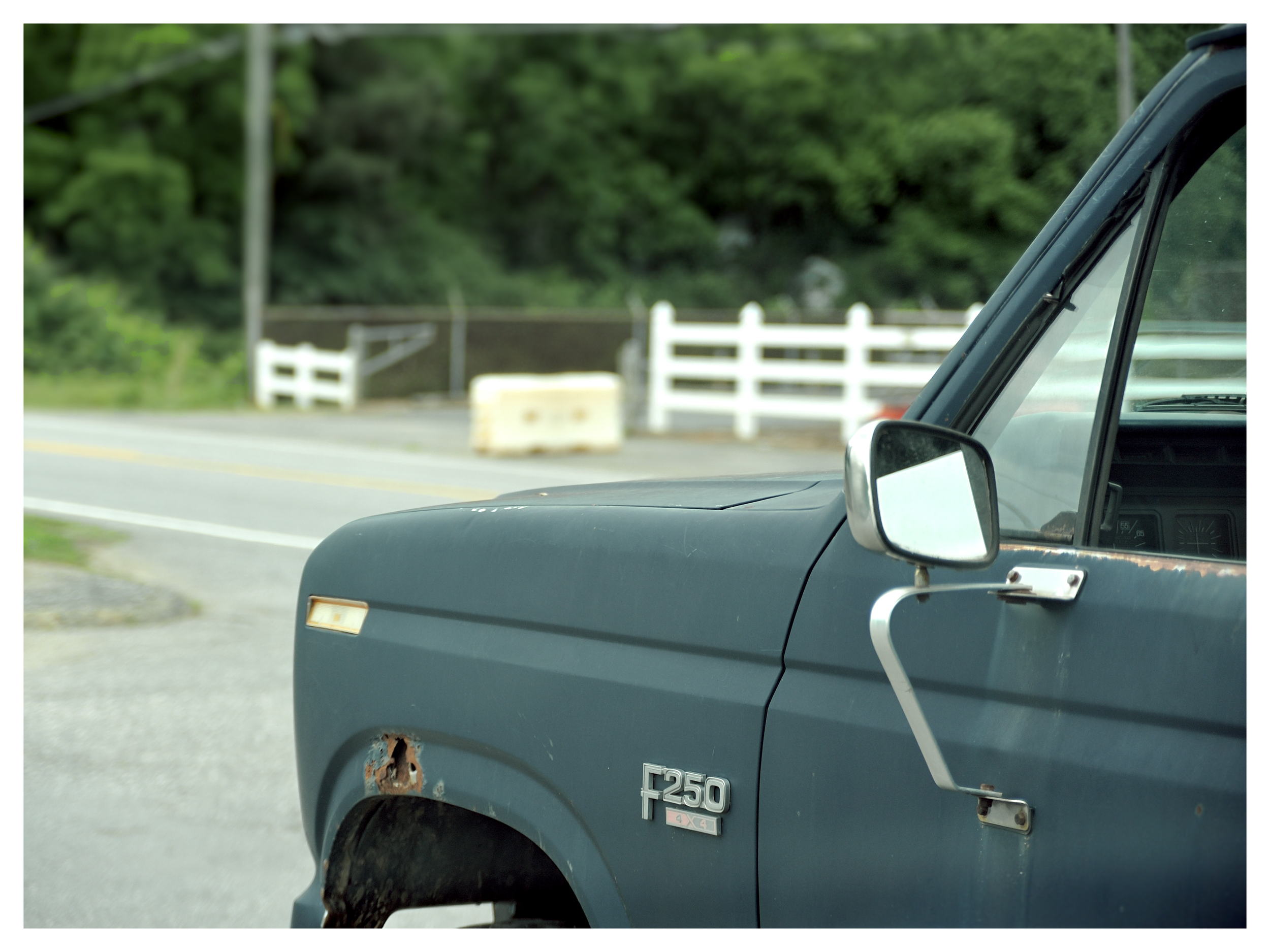 Close-up of the front right side of a black Ford F-250 pickup truck with rust damage near the wheel, parked near a white wooden fence and greenery in the background.