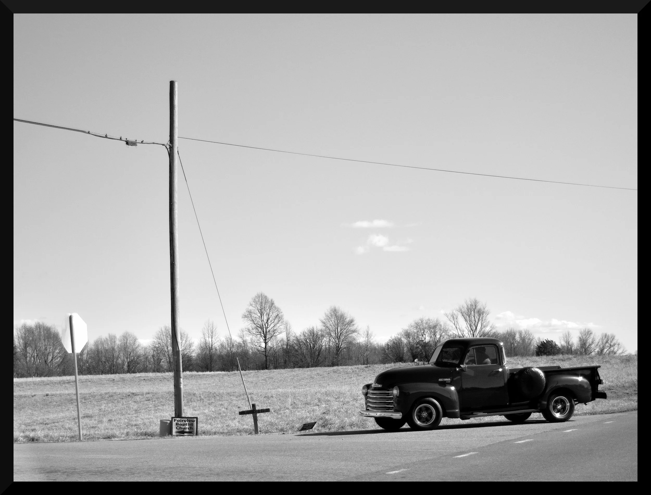 A classic small town image. Vintage truck, small 2 lane rd with crosses on the side of the road. There is a small roadside sign for Fairview Baptist Church.