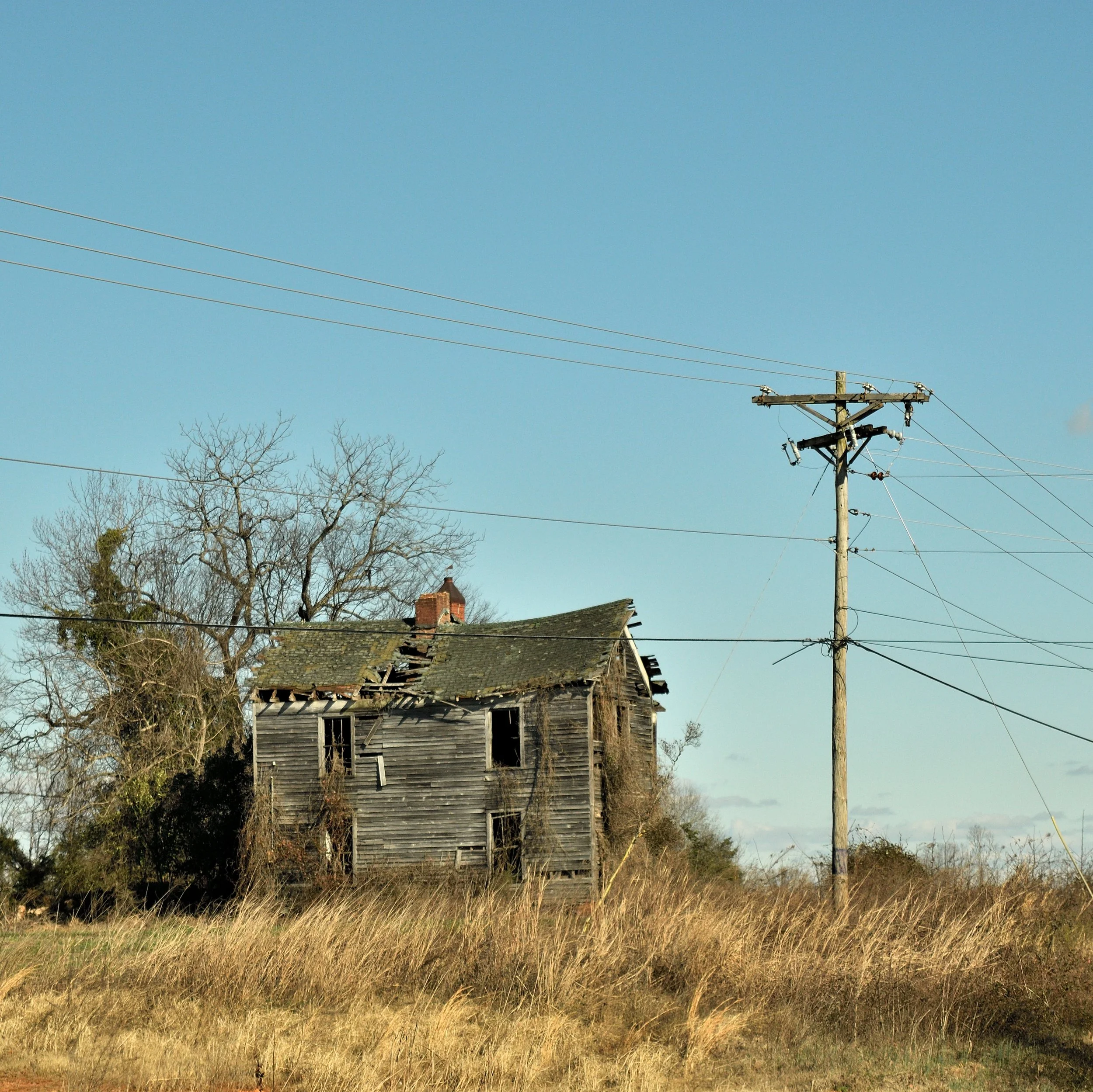 An old, dilapidated wooden house with broken windows, surrounded by tall grass and leafless trees, with a utility pole and wires in the foreground under a clear blue sky.
