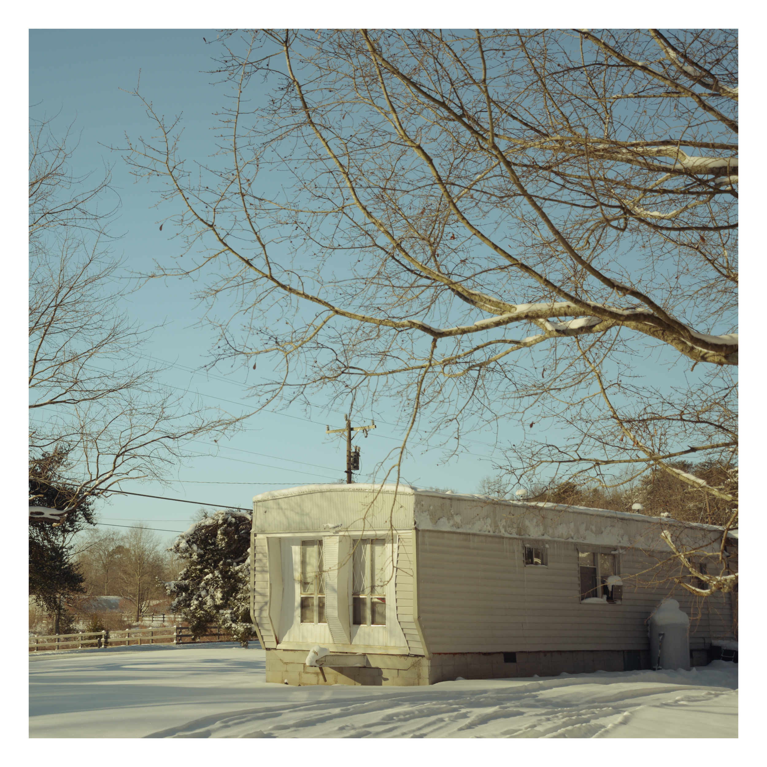 A snow-covered backyard with a white house, snow on the ground, and leafless trees against a clear blue sky.