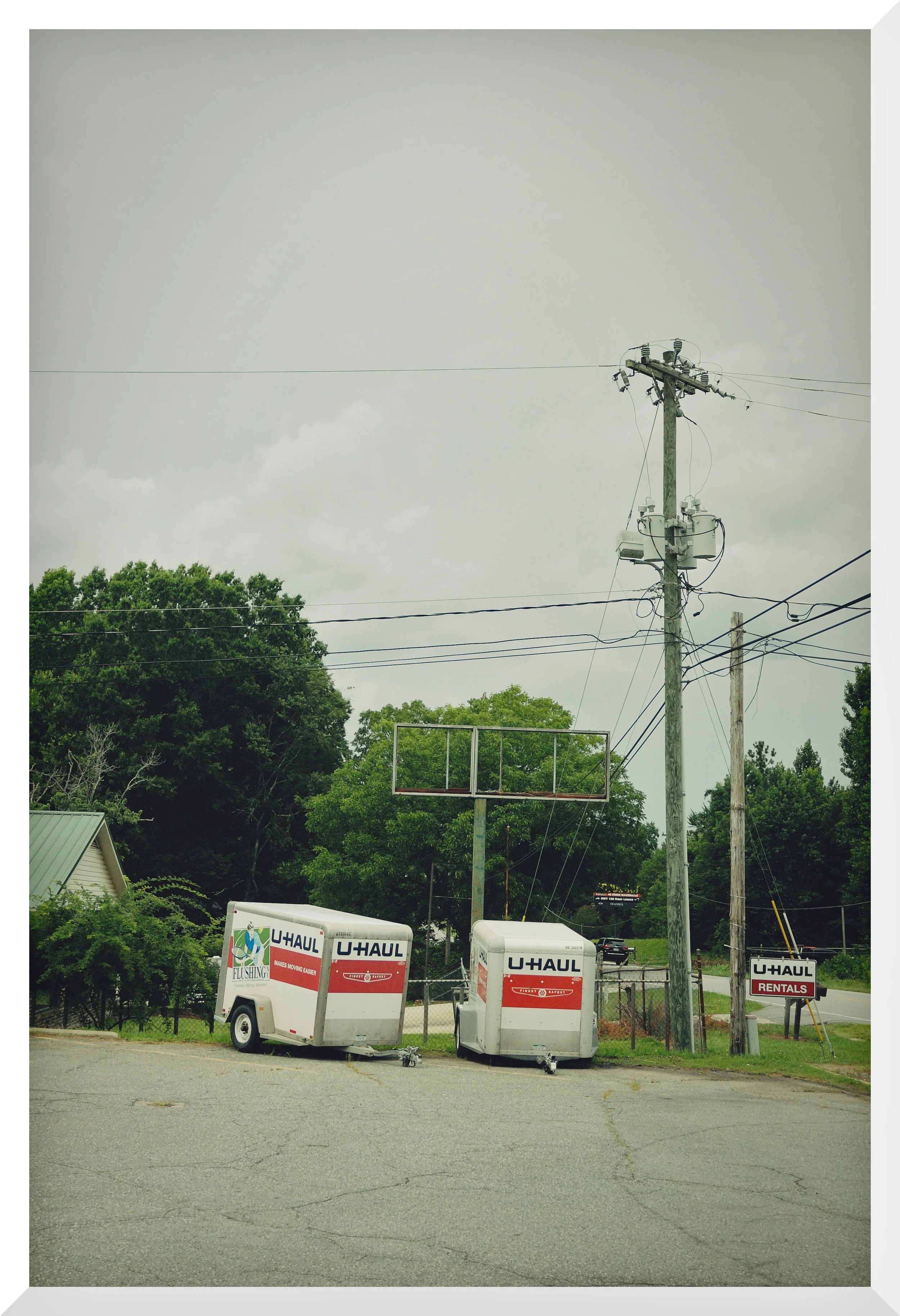 Several U-Haul rental trailers are parked on a paved lot in front of a green tree line. A utility pole with transformers and wires stands to the right. Two U-Haul trucks are visible in the background.