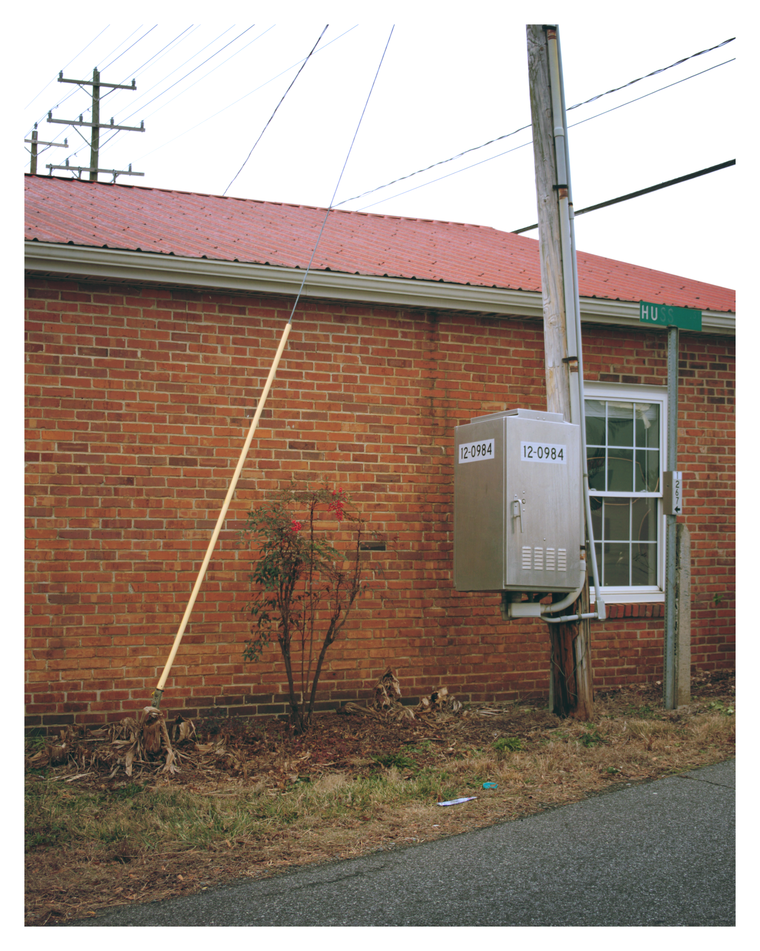 A brick building with a red corrugated metal roof. A utility pole with multiple electrical wires and a street sign reading 'Huss' are in front of the building. There is a small tree and some dried plants at the base of the building, and a utility box