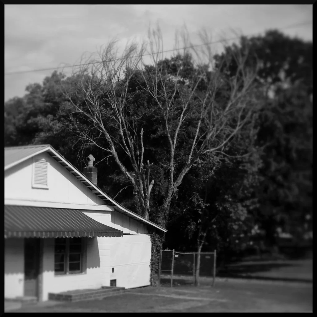 A black and white photo of a house with a metal roof, a window, a chimney, and a statue on top of the chimney. There are leafless trees behind the house and some taller trees in the background.