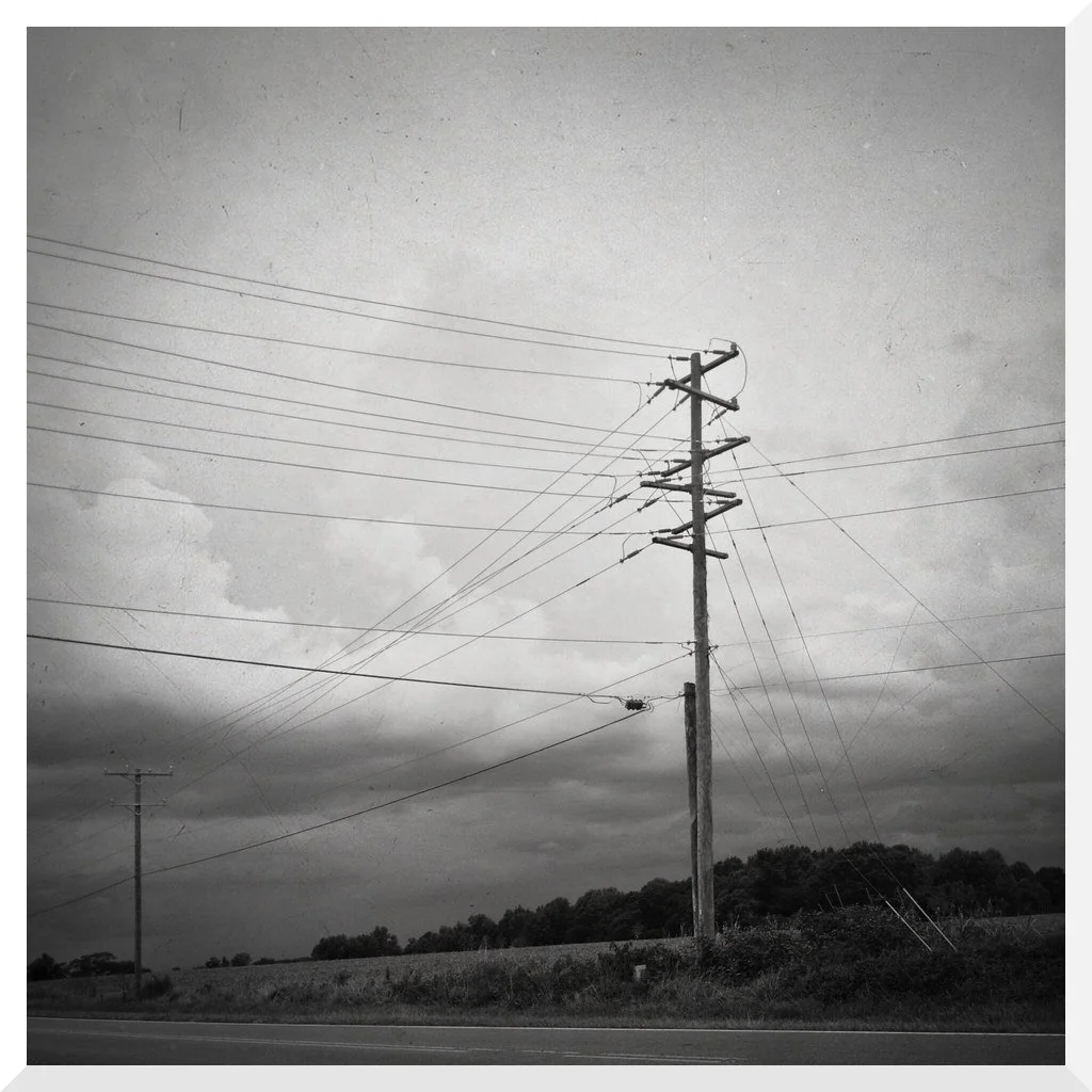 Black and white photograph of a rural scene featuring wooden utility poles with multiple power lines, overcast sky, and a tree-covered hill in the background.