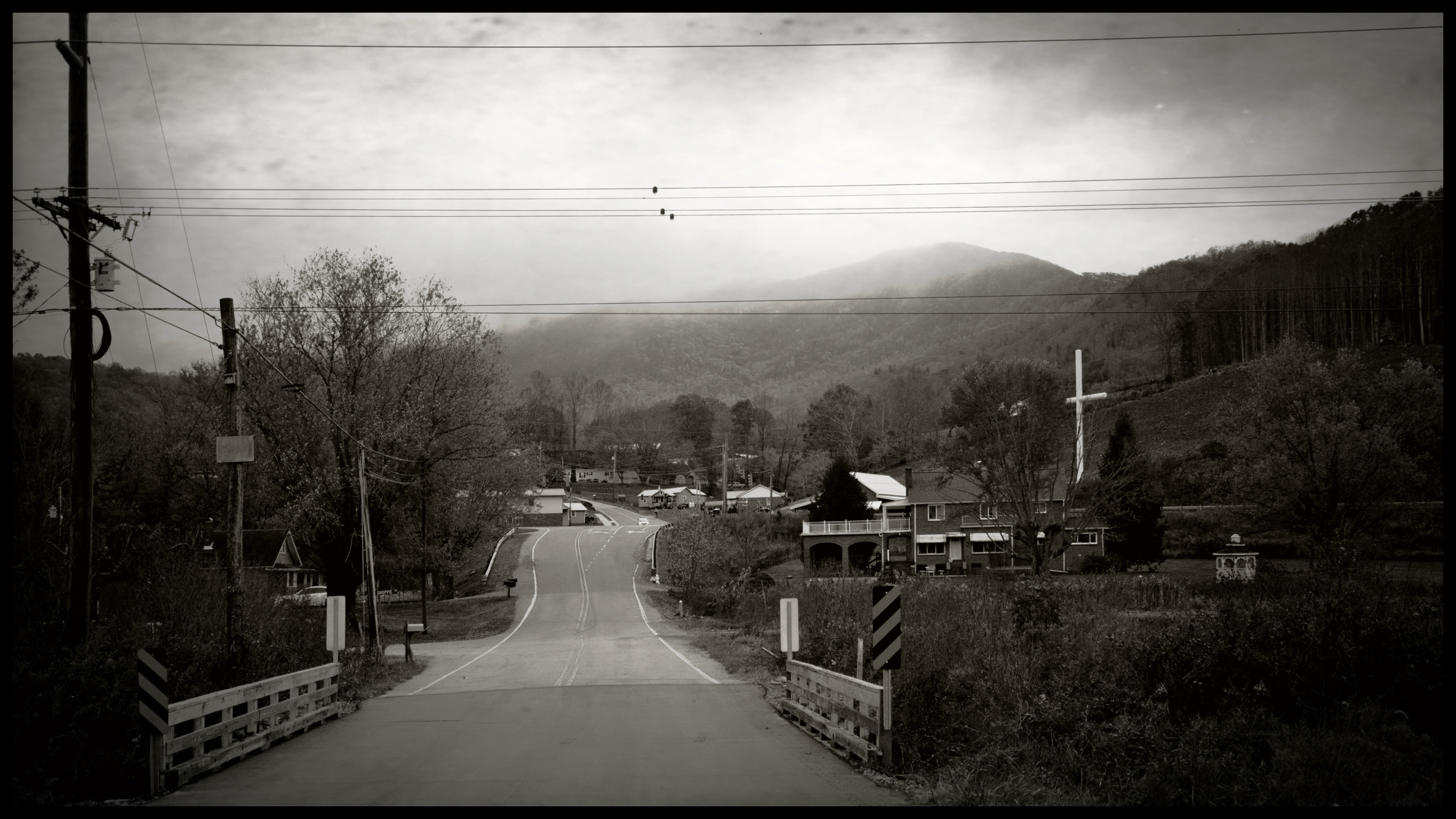 A black and white photo of a rural road with a small bridge, leading to a small town with houses, surrounded by trees and mountains in the background, under a cloudy sky.