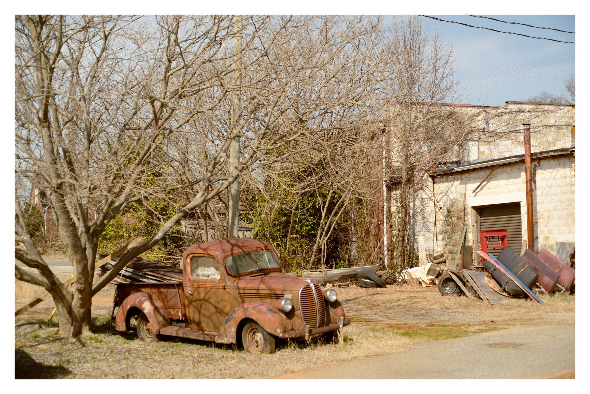 An old, rusted car parked next to a leafless tree in front of a weathered building with a partially open garage door, surrounded by various discarded objects and debris.