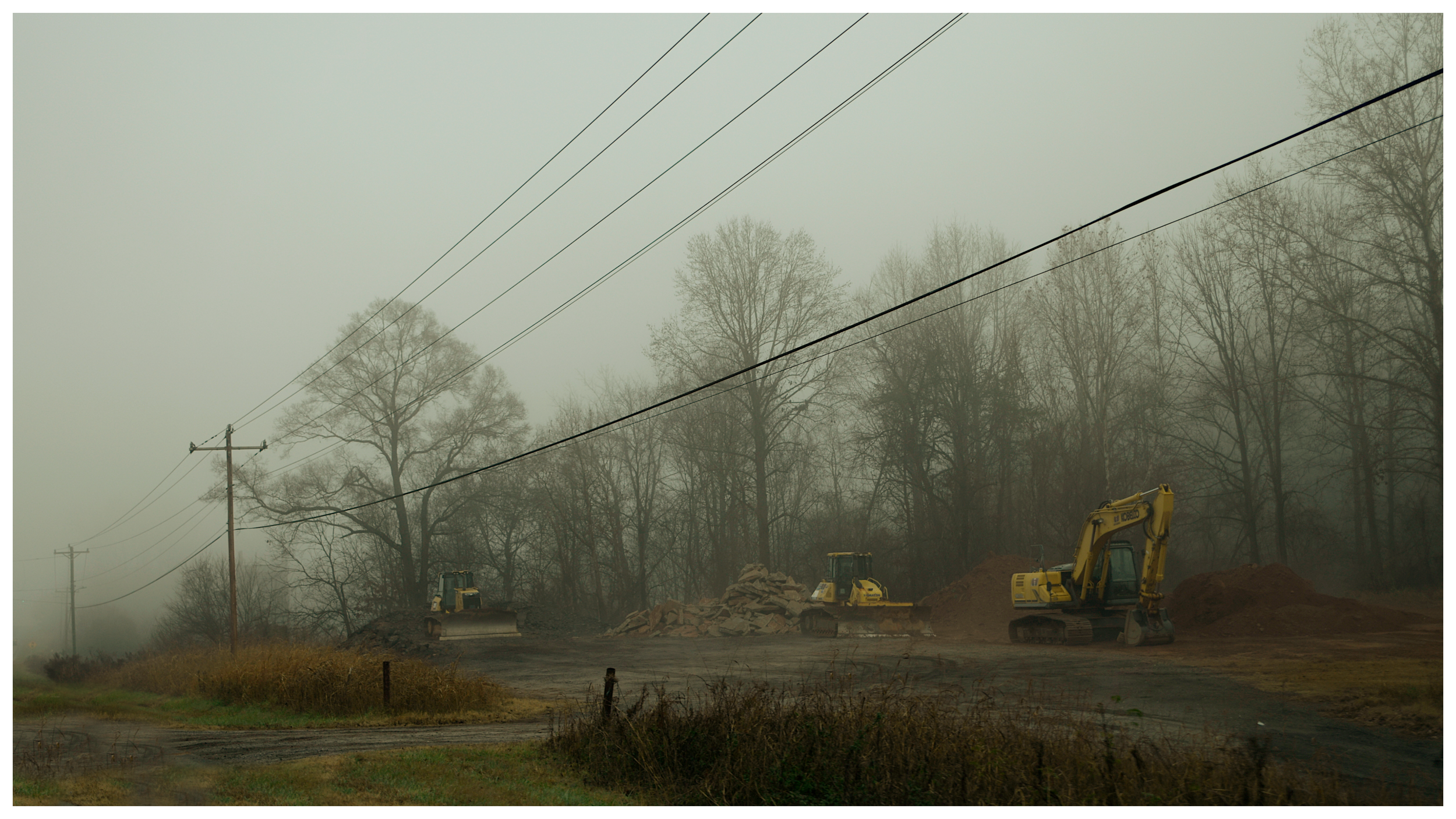 Construction site with excavators working on earth, surrounded by leafless trees and foggy weather.