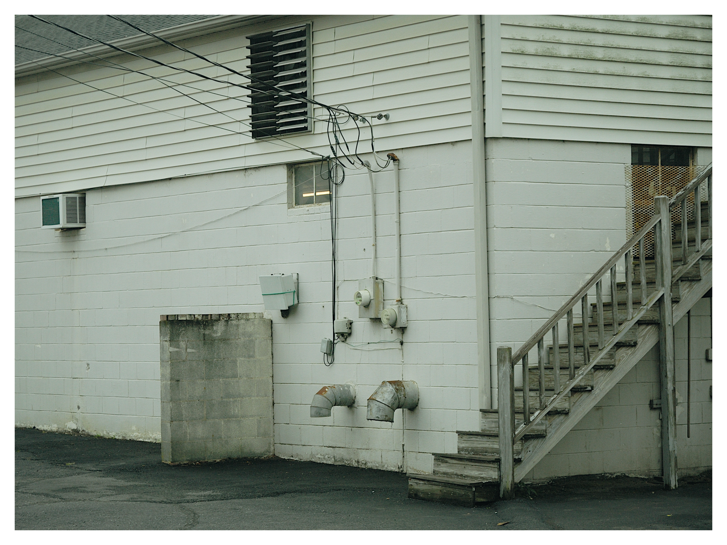 Back of an off-white building with horizontal siding, a small window, a window AC unit, several pipes, and electrical wires, with a wooden staircase on the right side.