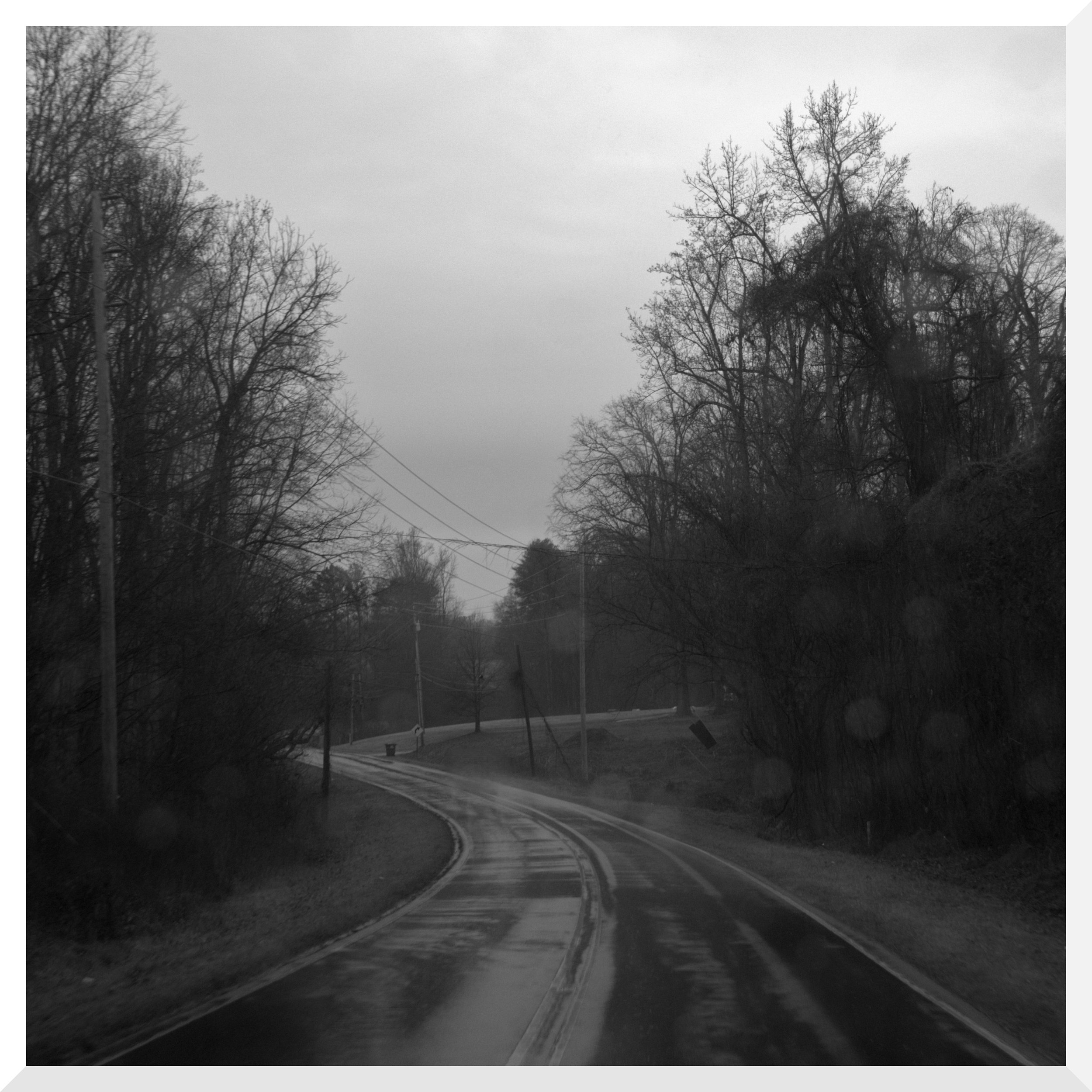 A black-and-white photograph of a winding residential street with bare trees, utility poles, and power lines under a cloudy sky.
