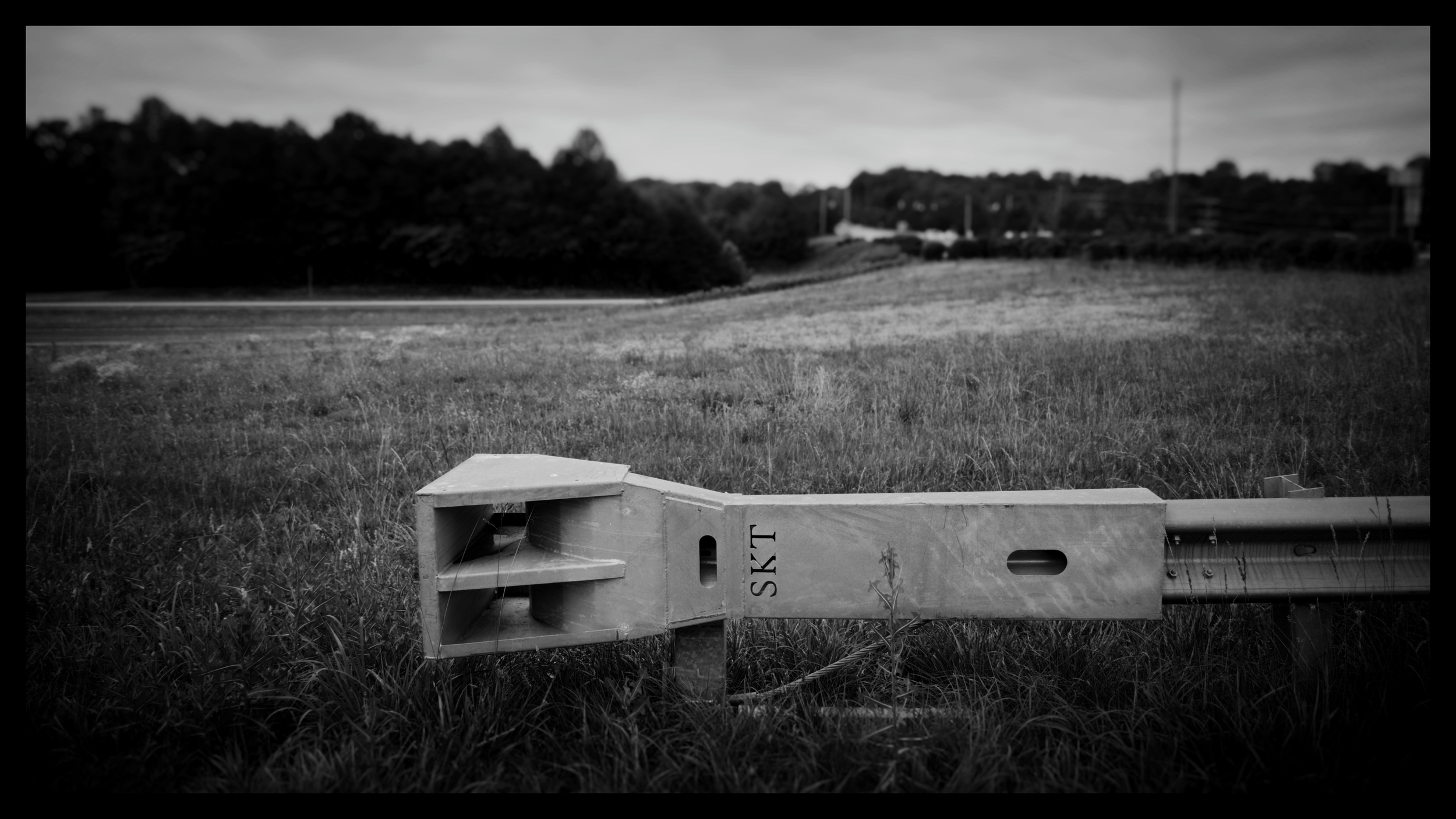 A black and white photo of a grassy area with a section of a metal or concrete structure in the foreground, with trees and power lines in the background under a cloudy sky.