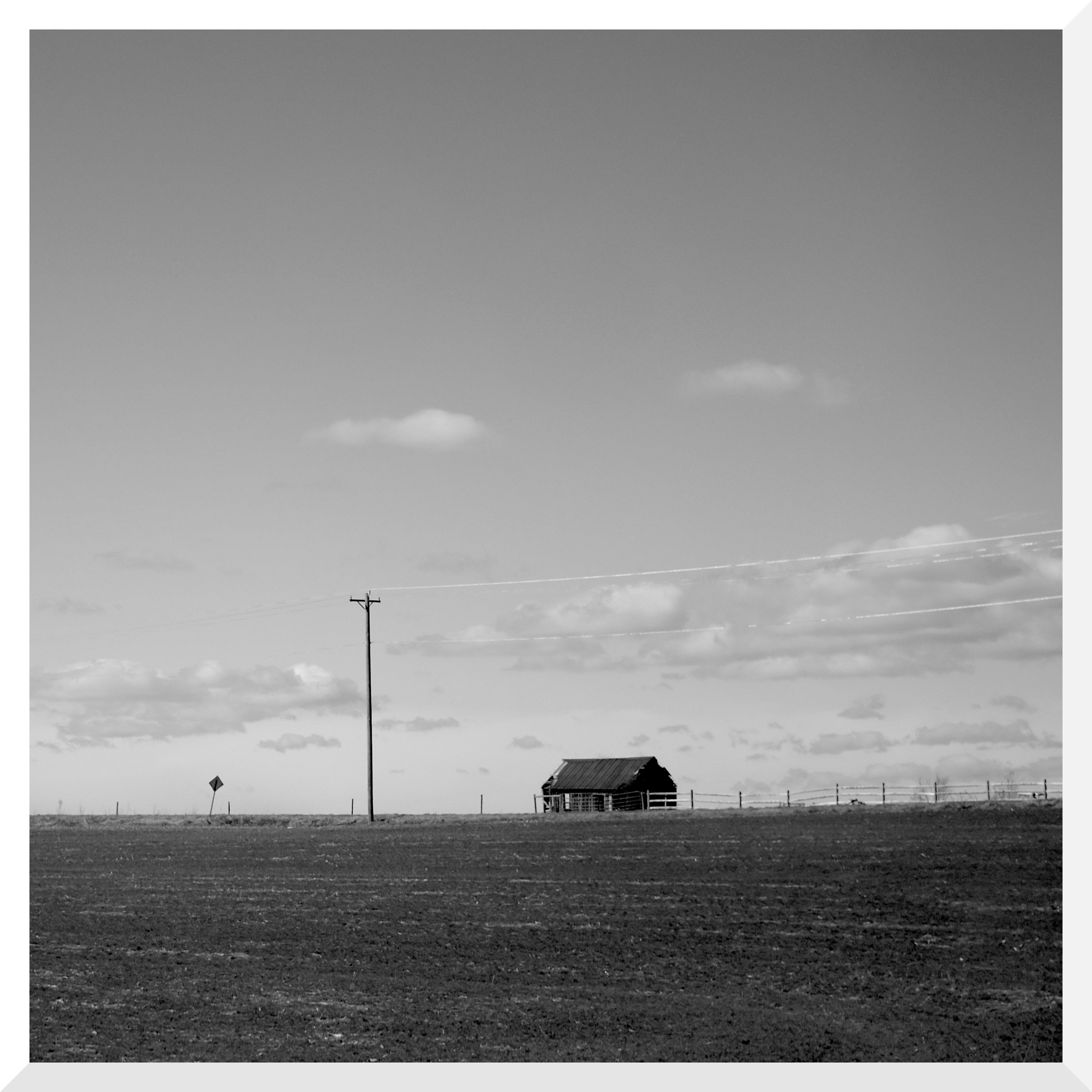 A black and white photo of a rural landscape with an open field, a power pole, and a small house or barn with a sloped roof against a sky with scattered clouds.